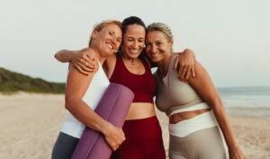 Three women standing together on a beach holding yoga mats, symbolizing shared support and wellness.