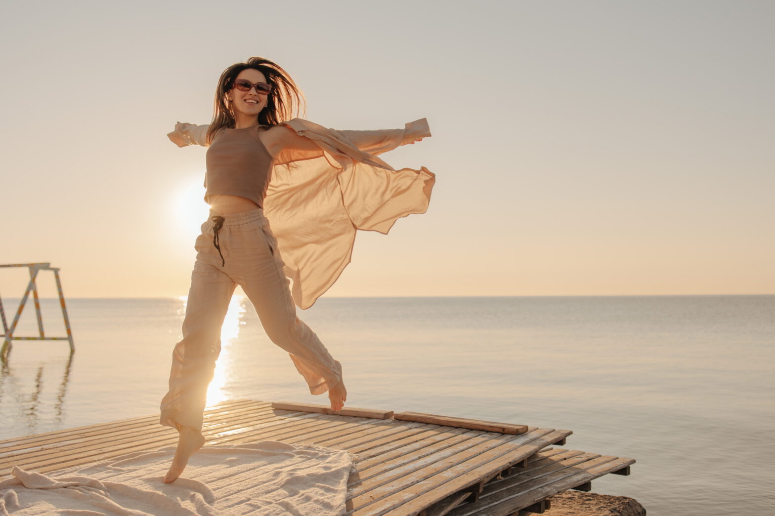 Woman joyfully jumping on a wooden dock at sunset by the water, wearing flowing clothing with the sun behind her.