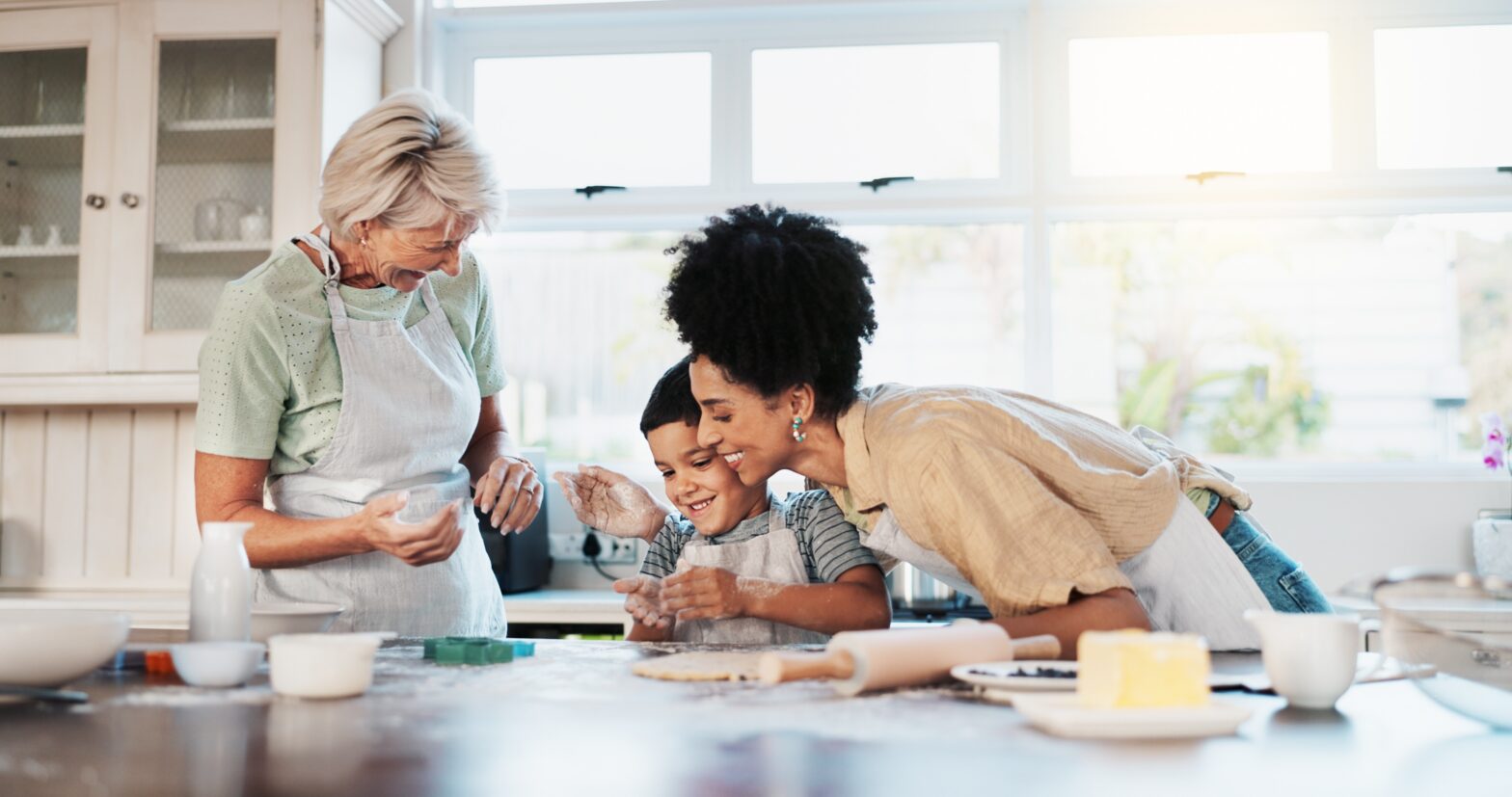 Grandmother, mother, and young child baking together in a bright kitchen, smiling and rolling dough on a countertop with baking ingredients spread out.