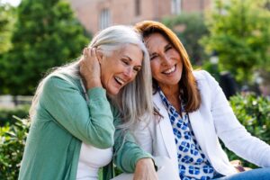 Two middle-aged women smiling and laughing together while sitting outdoors in a park setting.