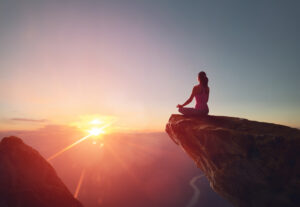 Woman sitting cross-legged in meditation on the edge of a cliff at sunrise, overlooking mountains and a valley below.