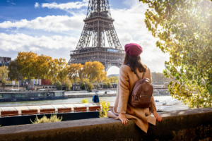 A person in a beige coat and red beret sits on a wall gazing at the Eiffel Tower in Paris