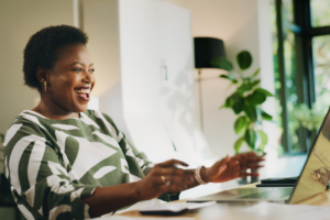 A woman smiles enthusiastically while looking at a laptop in a bright, modern workspace