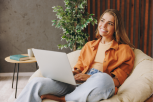 A smiling woman sits on a cozy chair using a laptop.