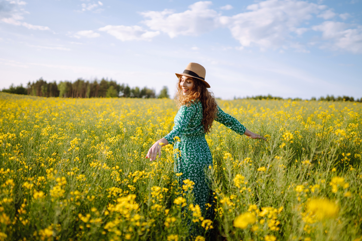 A woman with long curly hair wearing a green dress and a straw hat standing in a yellow flower field