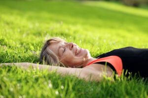 A woman who has a mindset for living a rich life lies on vibrant green grass, smiling with her arms stretched out, wearing a black and orange top.
