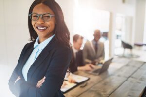 Smiling professional woman in glasses, arms crossed, stands confidently in a bright office.