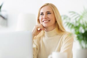 A smiling woman with long blonde hair sits at her financial planners desk, wearing a beige sweater.