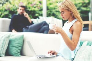 A financially independent woman with blonde hair sits on a white sofa, focused on her laptop while holding a cup.