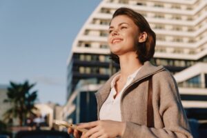 A successful woman outside of an office looking into the distance with her phone in her hand.